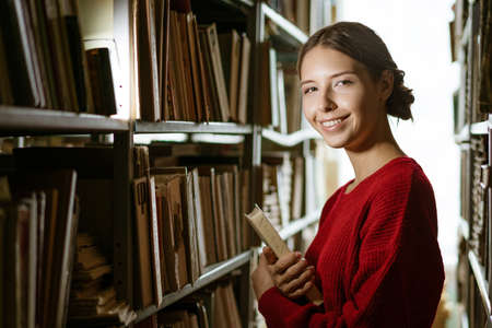 A young girl, dressed in a red sweater, holds a book in her hands against the background of the...