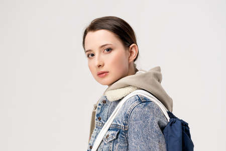 Young cute woman in a denim jacket and with a backpack posing on a white background.