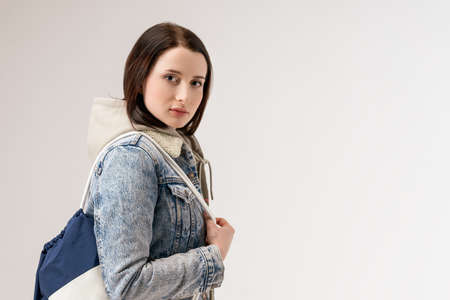 Young cute woman in a denim jacket and with a backpack posing on a white background.の写真素材