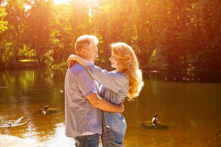 An adult couple stand on the shore of the lake in an embrace in the rays of the setting sun.の写真素材