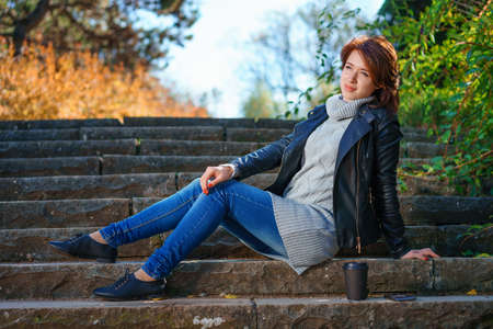 A young red-haired girl, dressed in a leather jacket with jeans, sits on the steps with a cup of coffee.の写真素材