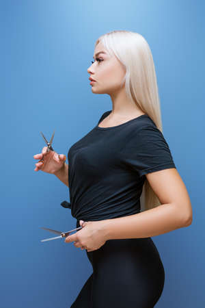 Beautiful blonde woman in black clothes, with scissors in her hands on a blue background, professional hairdresserの写真素材