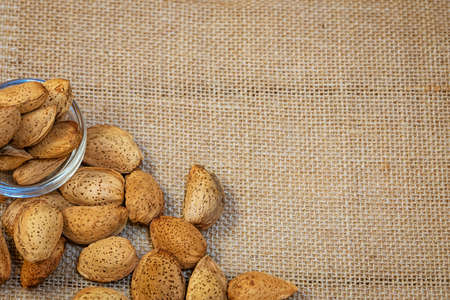 Raw almonds in the skin lies in a glass bowl and scattered on a sacking  background, close-up