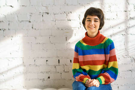 A young woman with short hair in a rainbow sweater and socks is sitting on a white sofa, the concept...
