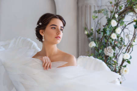 Portrait of a young beautiful woman wrapped in white angel wings. Cute girl romantic image on the background of a wall with columns and waving flowersの写真素材