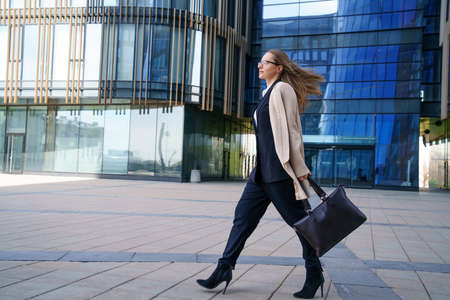 A business woman in a coat and suit, holding a bag in her hand, walks near the business center during the day. Conceptual horizontal photoの写真素材