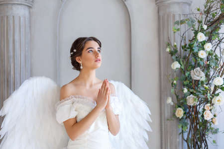 Portrait of a beautiful young woman in a white dress and angel wings, standing with a pleading look against the background of a wall with columns and flowersの写真素材