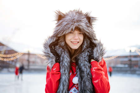Happy young woman in a wolf hat in winter at the ice rink poses in a red sweater outside in the afternoonの写真素材