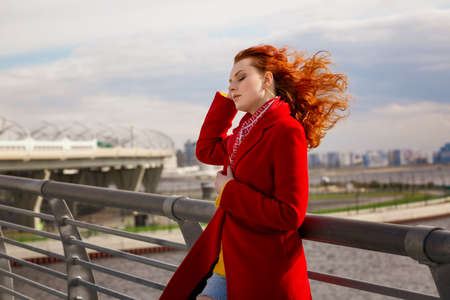 A woman in a red coat stands on the bridge, straightening her hair in the wind. Sunny day