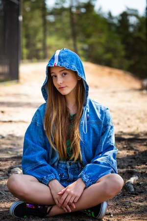 A young girl poses in a denim jacket with a hood against the background of a forest sittingの写真素材