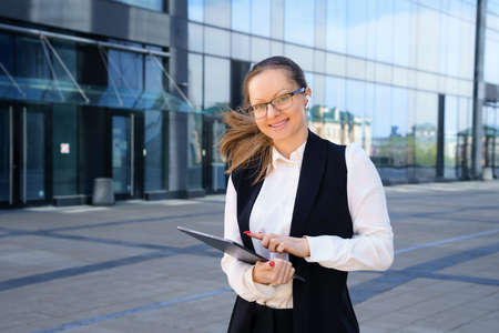 A business woman stands with a laptop in a suit and glasses outside an office building during the...