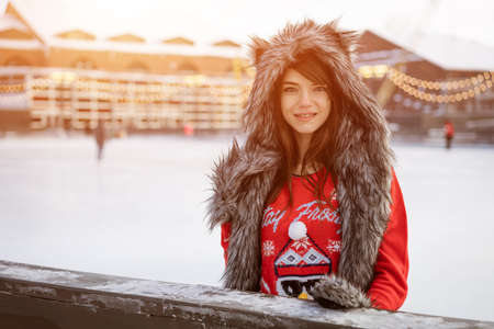 Happy young woman in a wolf hat in winter at the ice rink poses in a red sweater outside in the...