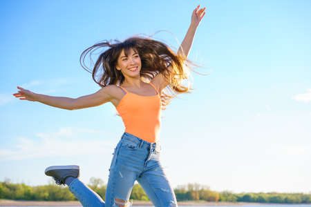 a young beautiful brunette girl in jeans and an orange t shirt jumps against the sky on a sunny...