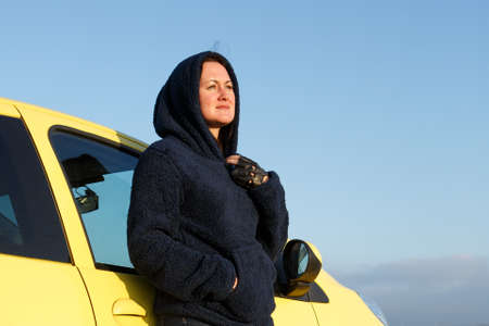 A woman looks away from a yellow car in a hood against a blue sky
