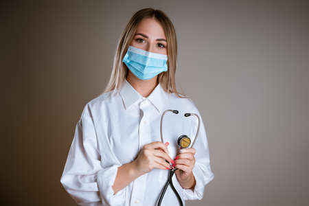 A young female doctor in uniform and wearing a mask stands against a gray backgroundの写真素材
