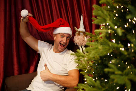Funny man in a Santa Claus hat shows a like gesture while sitting in a chair near the Christmas treeの写真素材