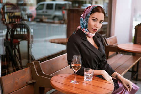 A beautiful brunette woman with bright makeup is sitting in a cafe on the street with a glass of...