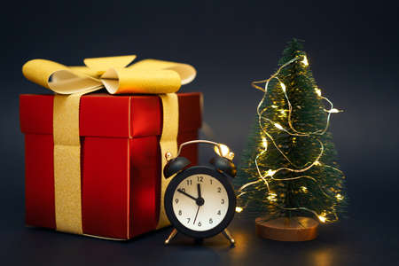 Red gift box with a gold bow. White alarm clock and a small Christmas tree on a black background.