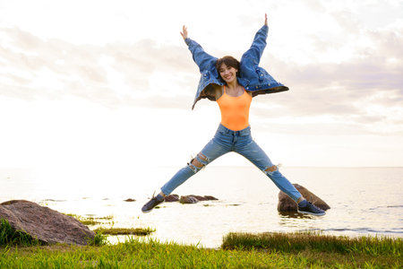 A young girl with long hair in jeans and a yellow t-shirt is jumping on a beautiful sunsetの写真素材