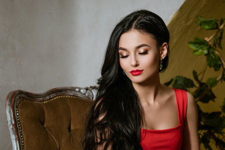 An elegant woman with long black hair in a red dress sits in a chair on a studio background. Luxury...