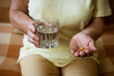 Mature woman with a glass of water holds a tablet in her hand while sitting on the couch. Treatment conceptの写真素材