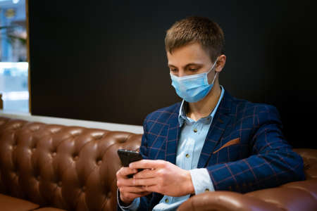 young business man in a medical mask sitting on the couch with a phone in his hand readingの写真素材