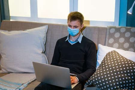 young caucasian man sitting at home on sofa with laptop in medical mask
