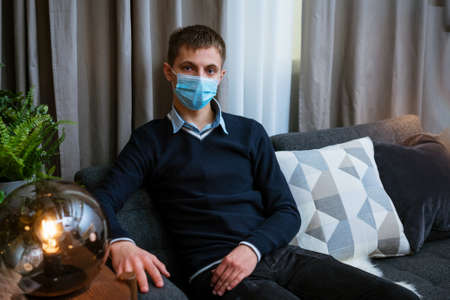 a young man in a medical mask sits on the sofa at home with a pensive lookの写真素材