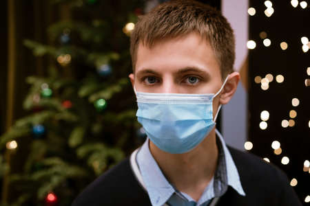 portrait of a young man in a medical mask and a sweater against a background of bokeh and a Christmas treeの写真素材
