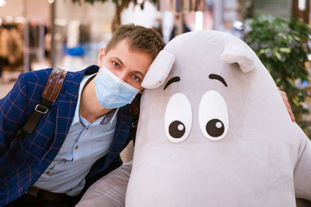 a man sits in a jacket and a medical mask in a mall near a soft toy