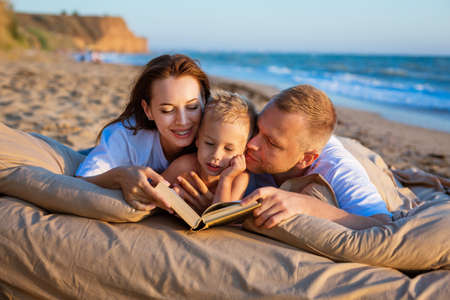 A young couple, dressed in light-colored clothes, lies with a child on the seashore and read a book. Relaxation. Family concept. Everyday leisure.の写真素材