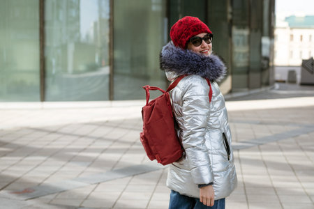 Happy young woman in sunglasses in a winter jacket with a backpack near the building. Travel conceptの写真素材