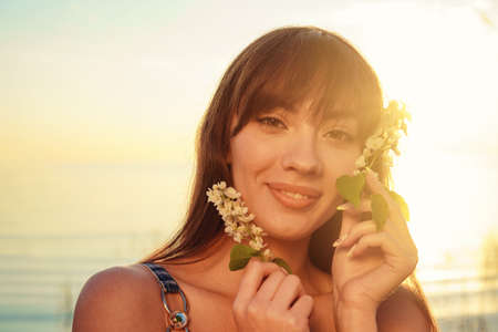 Portrait of a cheerful European young woman against a background of water, holding flowers near her face. Natural cosmetics conceptの写真素材