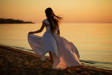 Young free woman running in white dress near the sea at sunsetの写真素材