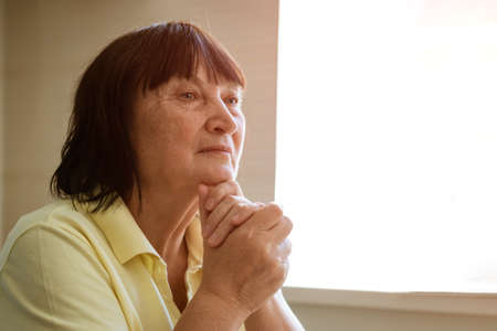 A mature woman sits at a table in the kitchen by the window with a pensive look