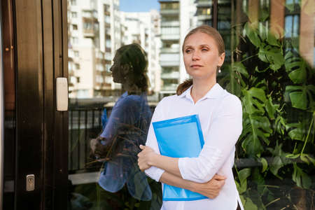Business successful woman in white shirt with blue folder for papers stands at office building