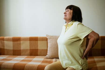 Mature woman holding her back with her hand in pain while sitting on the sofaの写真素材