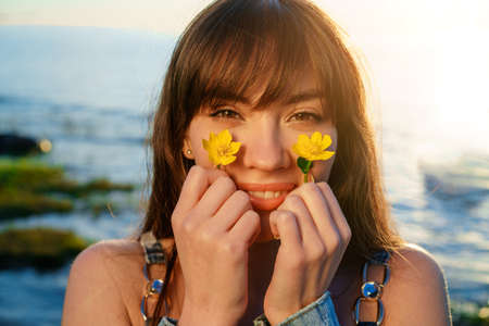 Portrait of a cheerful European young woman against a background of water, holding flowers near her...