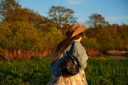 Young woman in a hat with a backpack in jeans outdoors at sunsetの写真素材