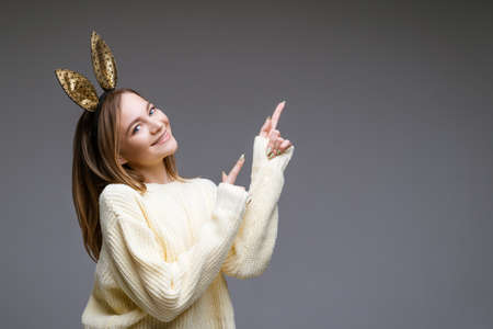 Portrait of a beautiful cheerful young woman in a sweater and in bunny ears shows her finger to the...