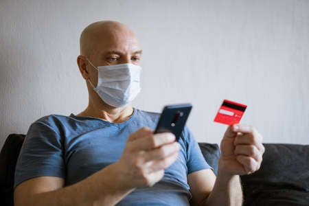 Bald man in protective mask with phone and credit card in hand, internet shopping concept