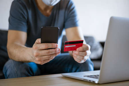 Bald man in a medical mask with a credit card sits on the couch at a laptop with a phone in his...