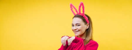 smiling young caucasian woman, blonde with pink bunny ears, shows a heart with two hands and looking at camera isolated over yellow backgroundの写真素材