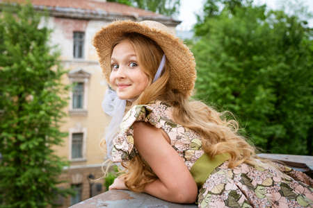 Little beautiful girl in dress and hat posing on the balcony against the background of the city