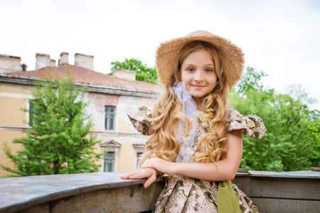 Little beautiful girl in dress and hat posing on the balcony against the background of the city