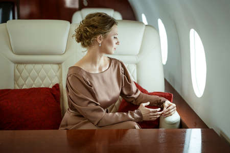 an attractive blonde woman in an elegant beige dress sits in a comfortable seat on a private jetの写真素材