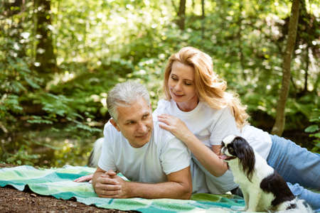 happy mature married couple in forest lie on blanket with dog, picnic conceptの写真素材