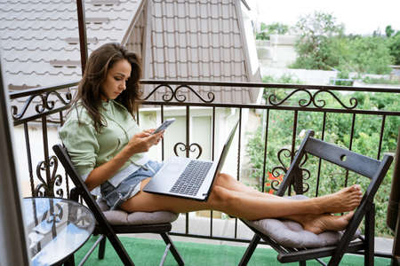A young woman in home clothes sits on the balcony, at a table works with a phone at a laptop in the summer afternoonの写真素材