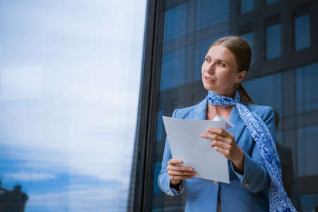 Business successful caucasian woman in blue jacket holds documents in hand near office buildingの写真素材