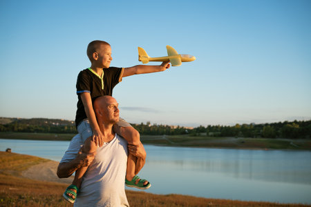 Father holds his son on his shoulders against the backdrop of nature by the lake at sunset. The boy...